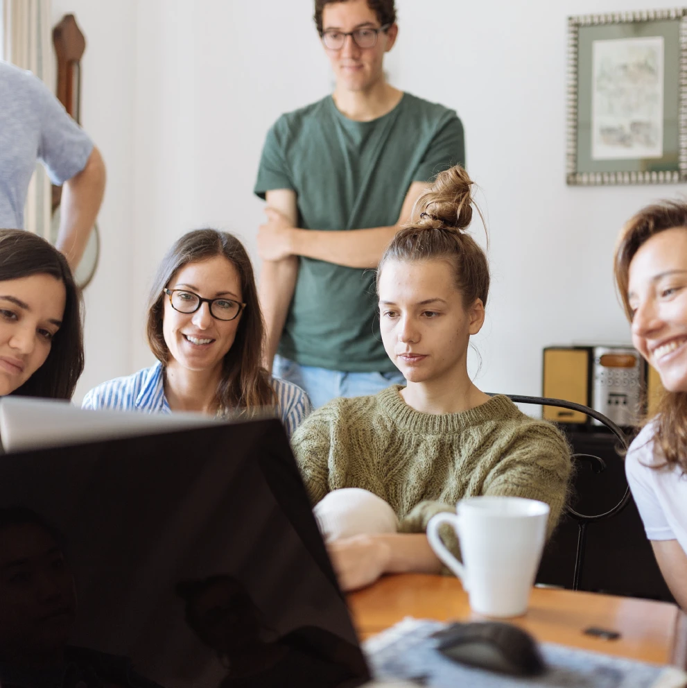 Graphic showing a group of five young professionals sitting around a table, collaborating and looking at a laptop screen together. One person stands behind them, and a large coffee mug sits on the table. The atmosphere is casual and friendly, suggesting a team meeting or remote work collaboration.