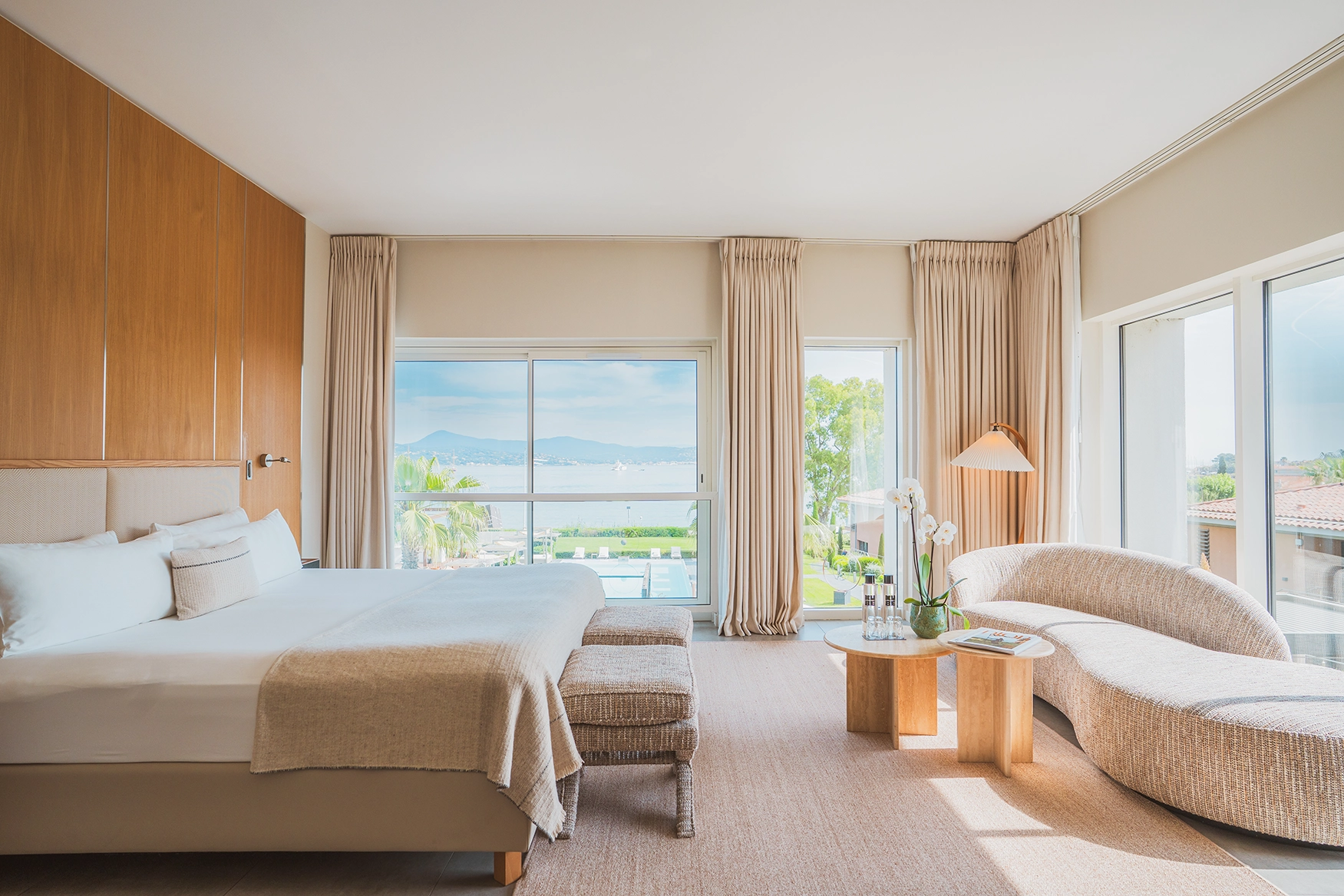 Modern hotel room with large bed, curved sofa, wood accents, and view of pool and sea through windows.