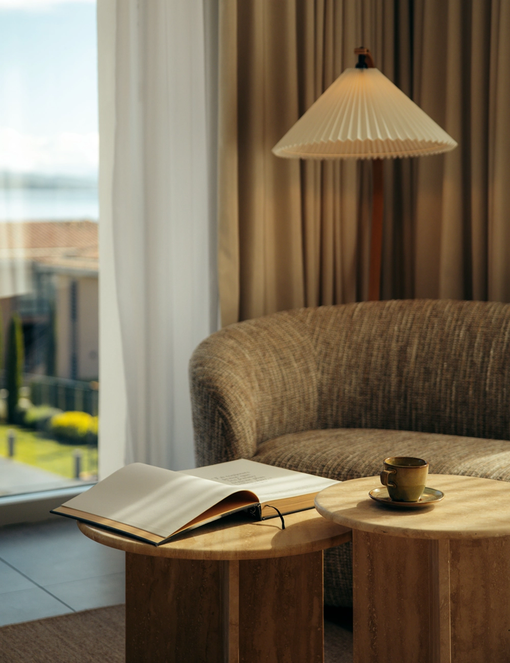 Cozy living room with a curved sofa, open book, cup on wooden tables, and a pleated lamp near curtained window.