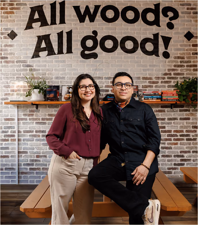 Debby and Fernando pose at 2Fifty BBQ beneath the “All wood? All good!” sign.