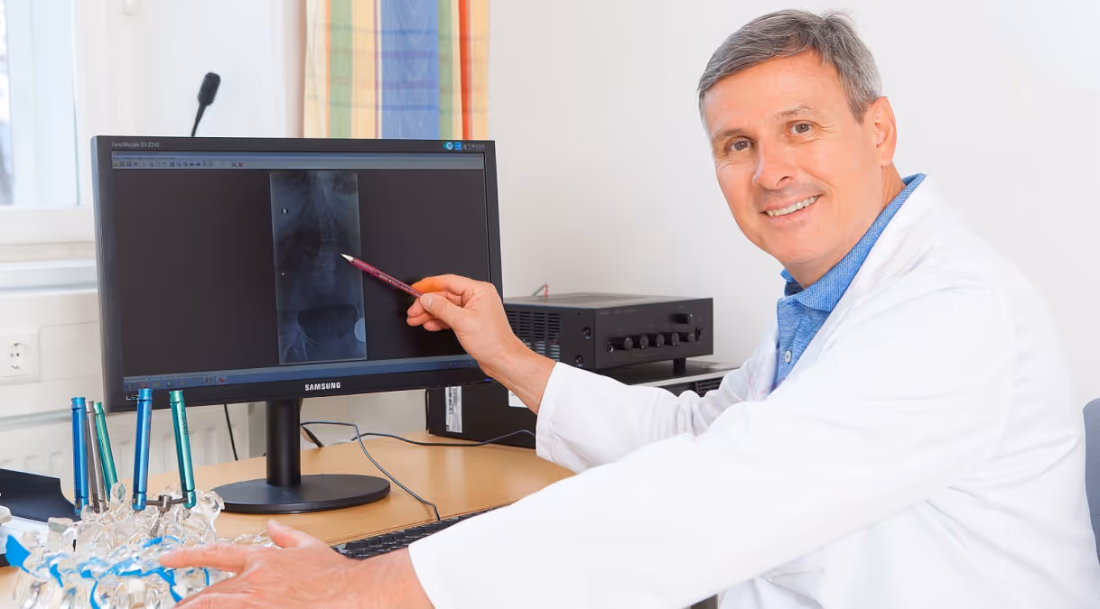 Facharzt Dr. Ronald Sabitzer zeigt auf Röntgenaufnahme der Wirbelsäule am Monitor und erklärt die Diagnose mit einem Wirbelsäulenmodell auf dem Schreibtisch