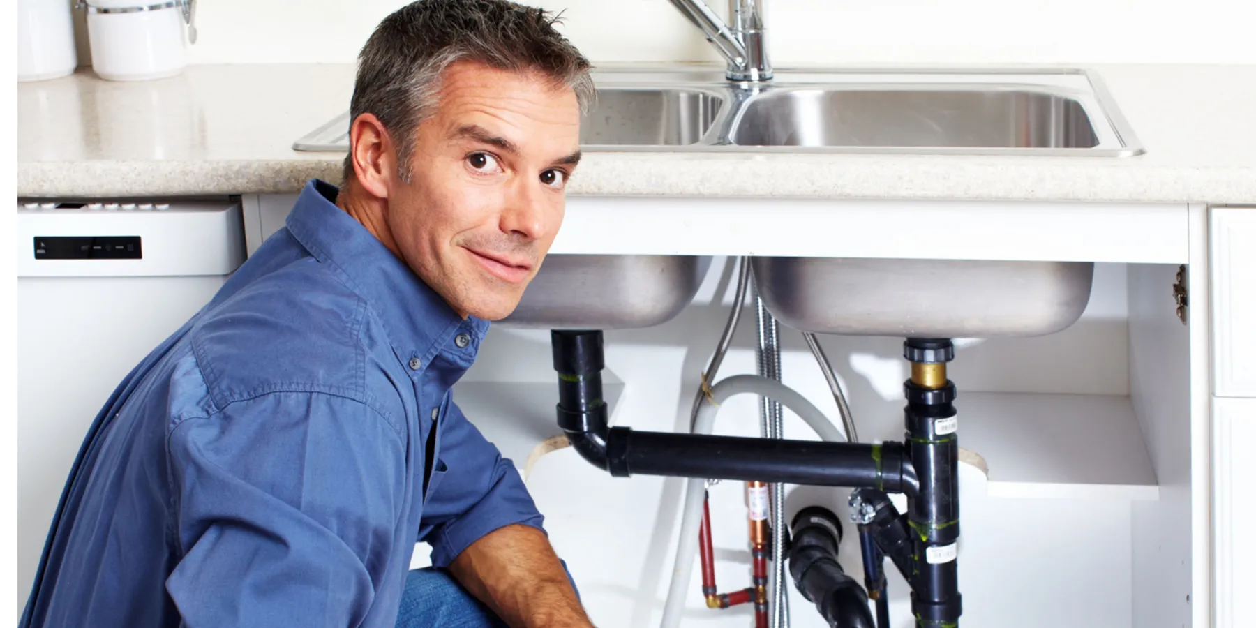 Man in blue shirt kneeling and inspecting plumbing under a kitchen sink.