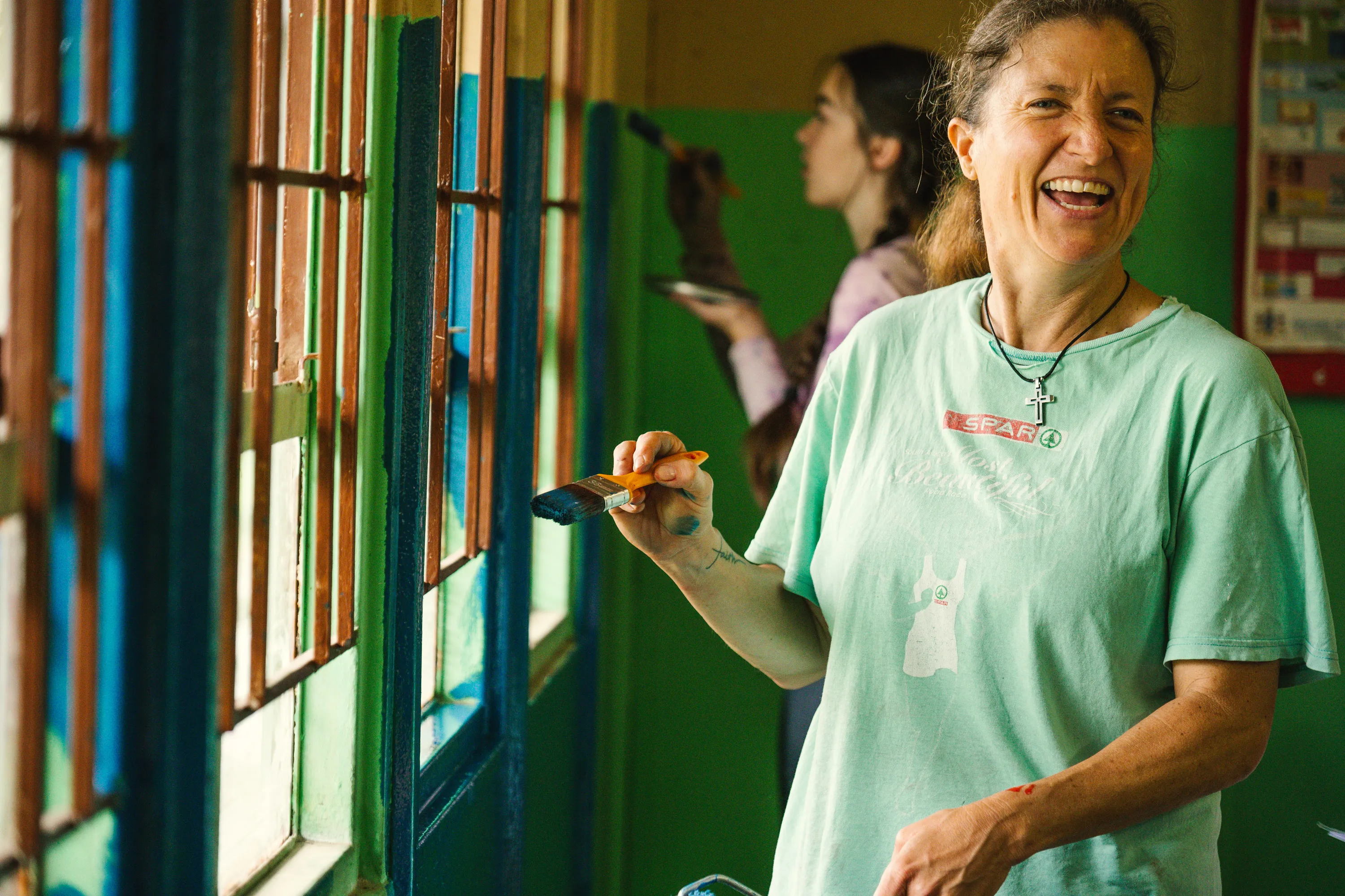 Volunteer smiling while another paints a windowsill in a classroom.