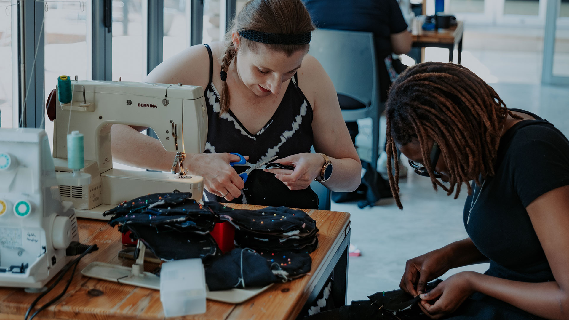 Two Love Our City volunteers stitching at sewing machines during a sewing event.