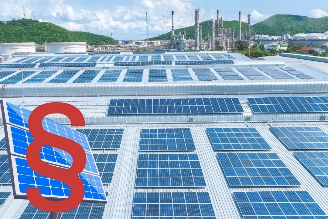 Auf dem Foto ist ein Gewerbedach zu sehen, auf dem Solaranlagen liegen. Im Hintergrund ist eine Fabrik und grüne Hügel zu sehen.