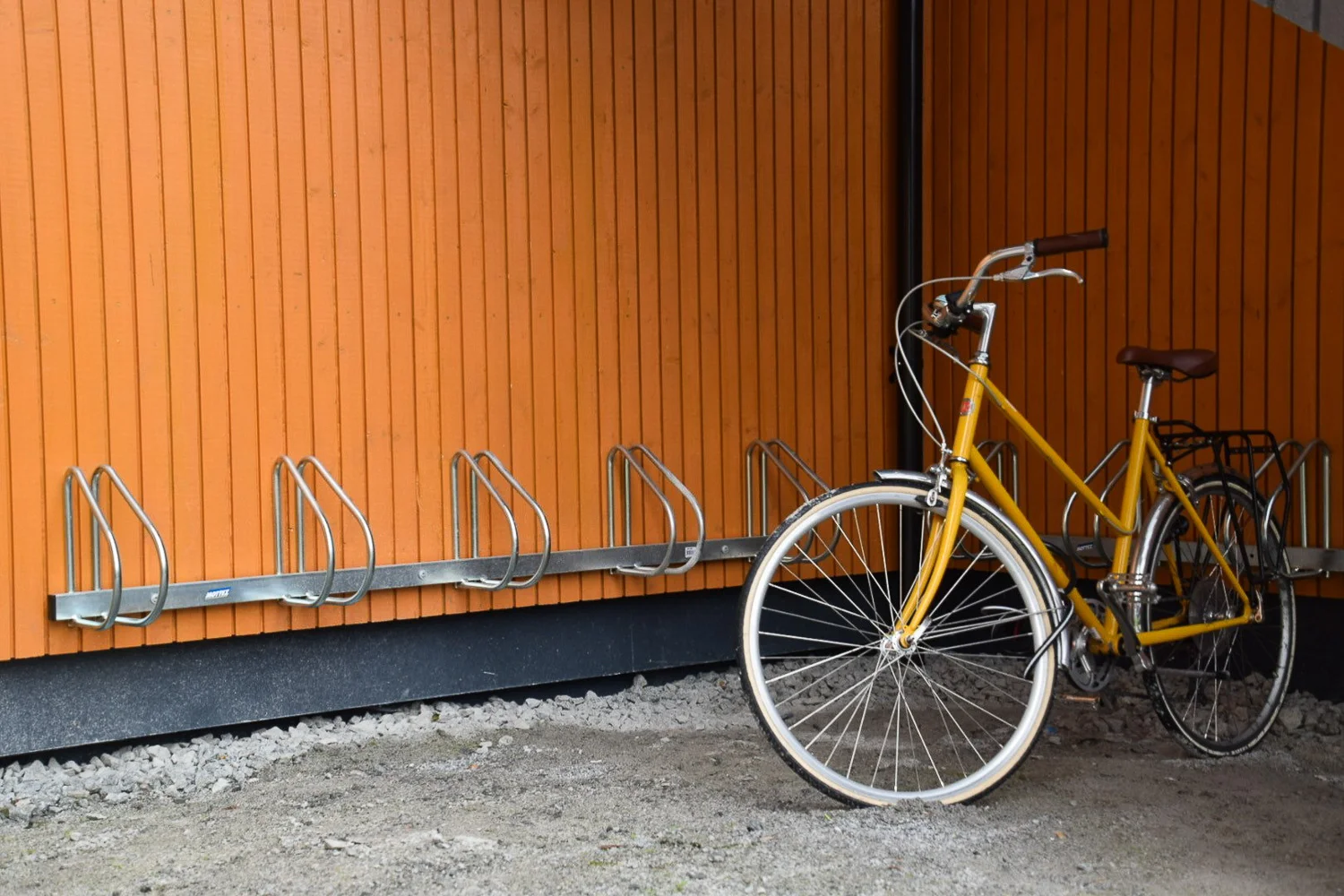 Yellow bicycle parked next to an empty metal bike rack against an orange wooden wall.