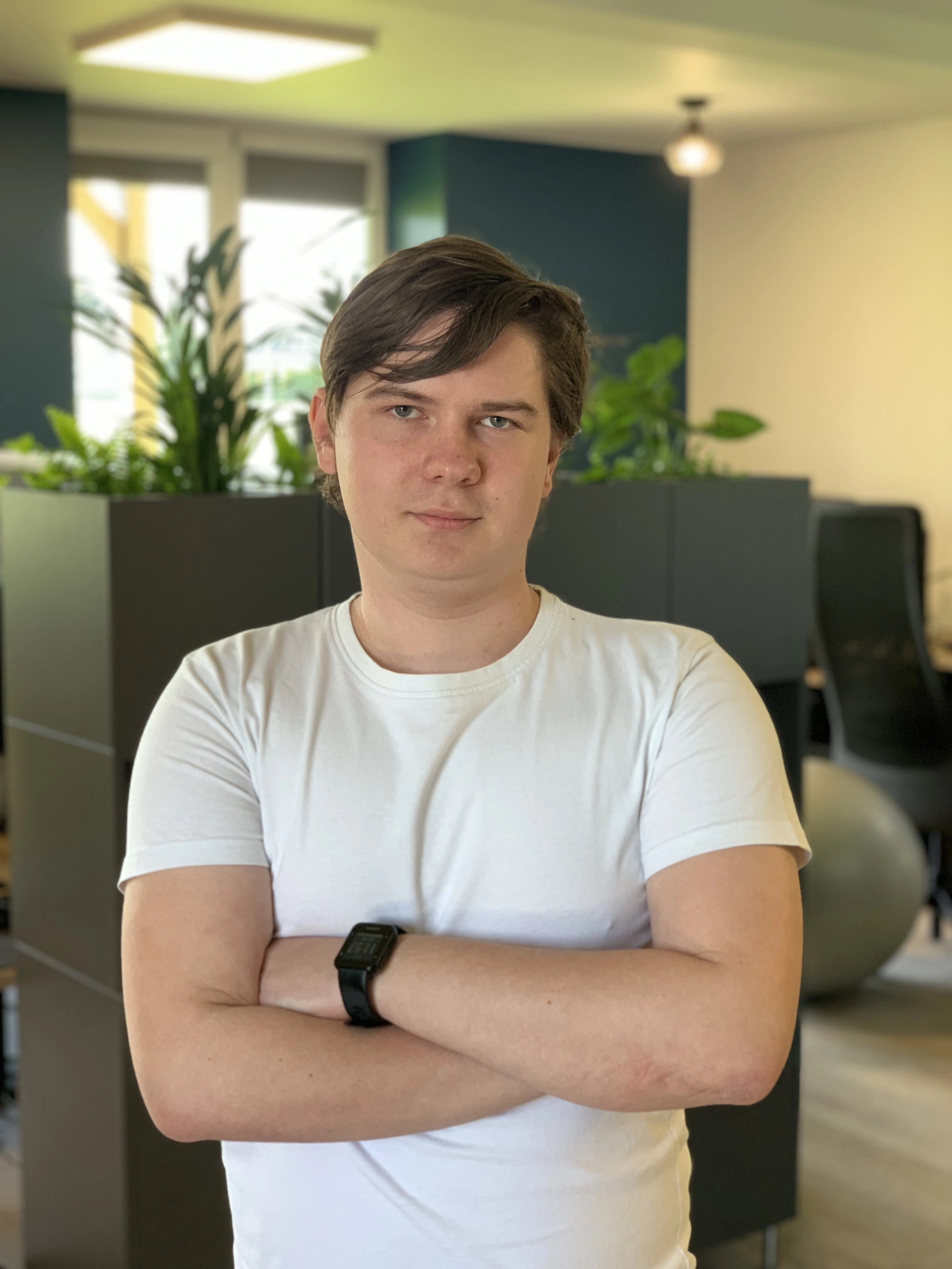 A person with short hair, wearing a white T-shirt and black watch, stands with arms crossed in an office with plants.