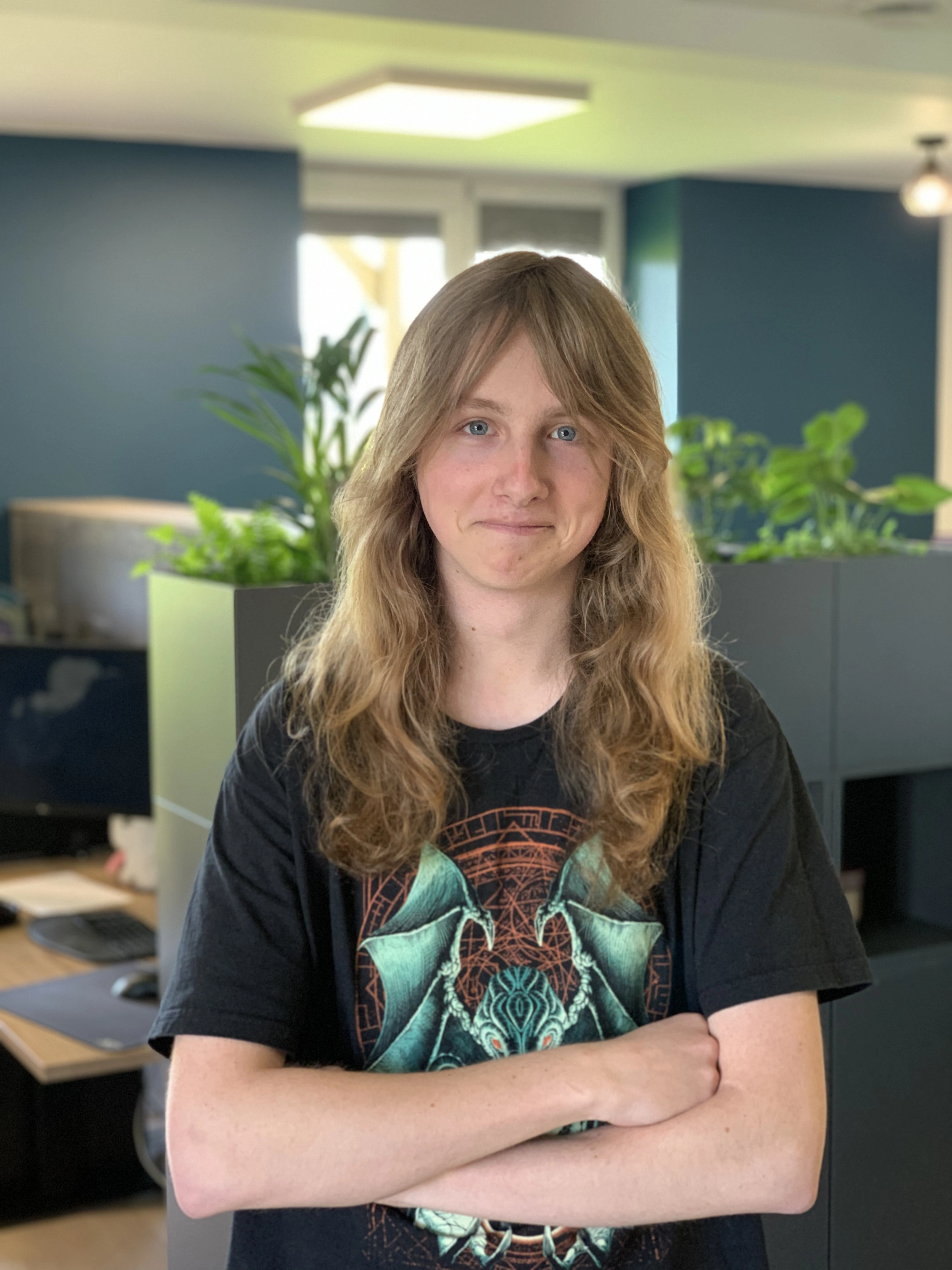 A person with long hair and a graphic T-shirt stands with arms crossed in an office with plants in the background.