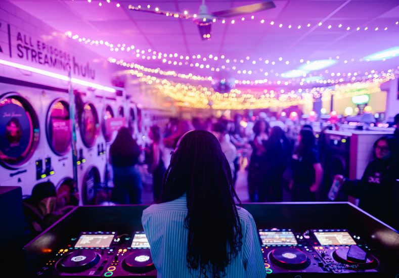 A DJ performs at a lively laundromat event, surrounded by washing machines, partygoers, and colorful string lights.