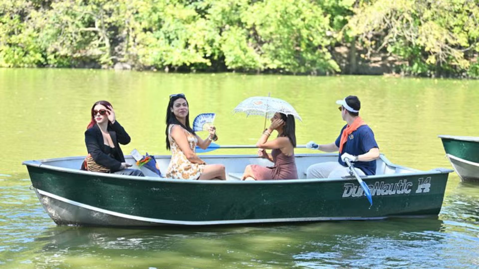 Four friends enjoy a peaceful day in a green rowboat on a calm lake, relaxing under a parasol with fans while one person rows.