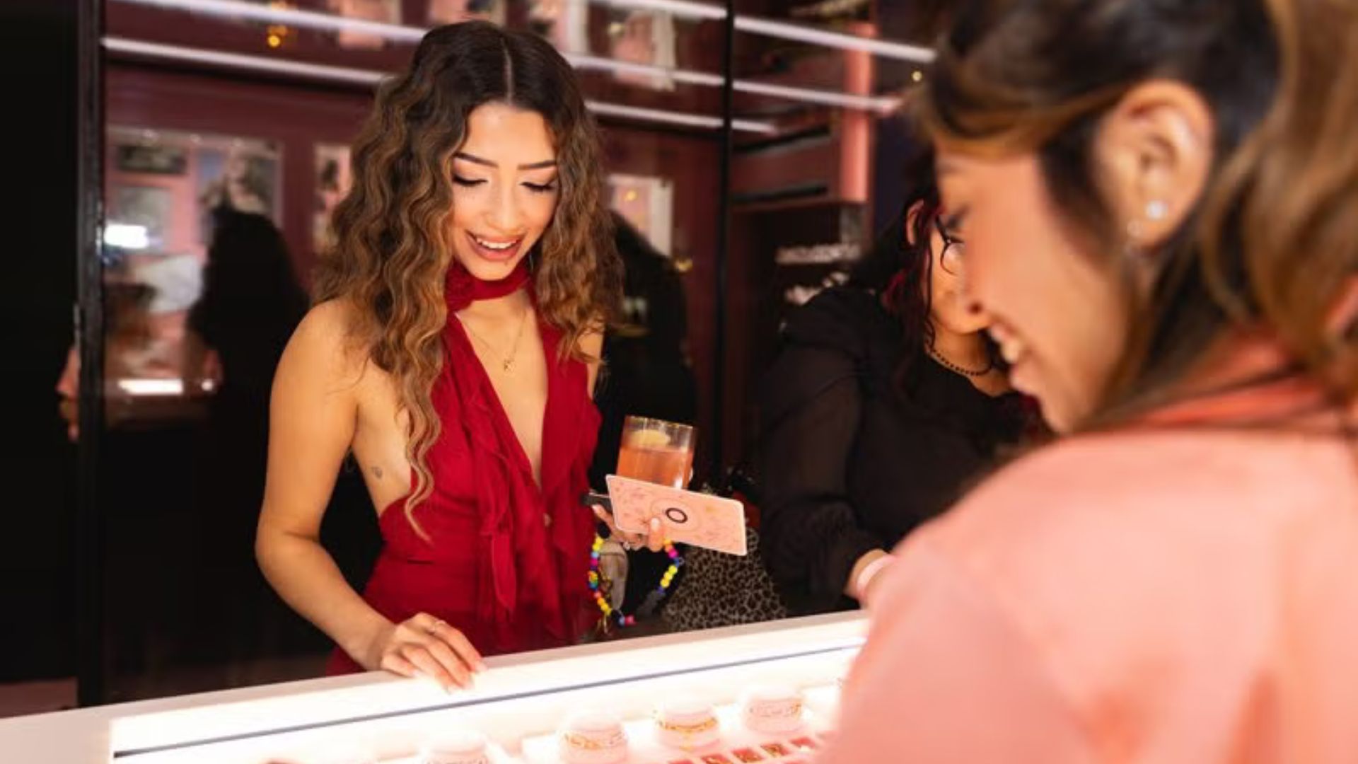 A woman in a red dress smiles while choosing charms at a brightly lit jewelery charm bar during a brand event.