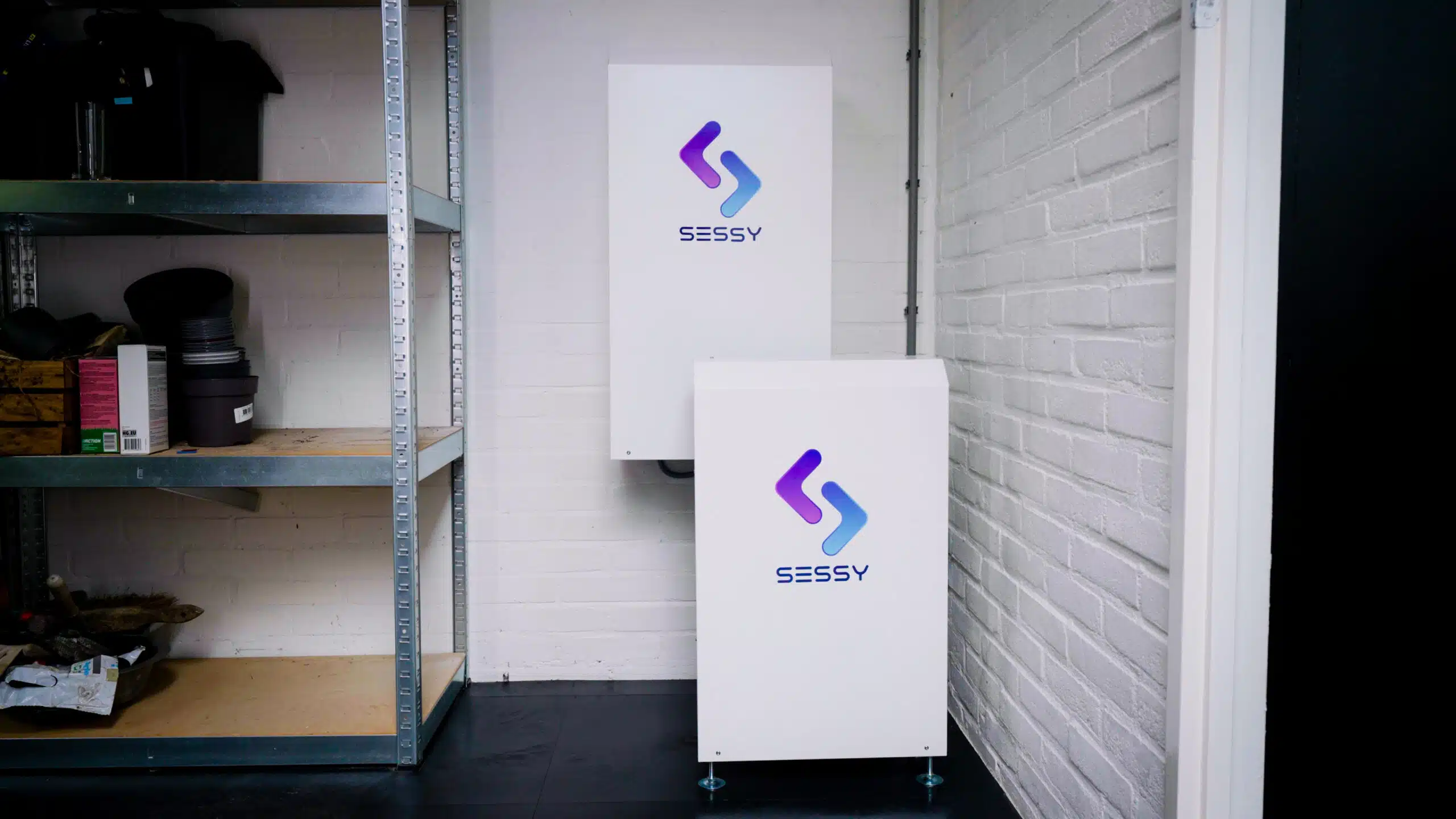 Witte thuisbatterij en omvormer met Sessy-logo geplaatst tegen een witte bakstenen muur.