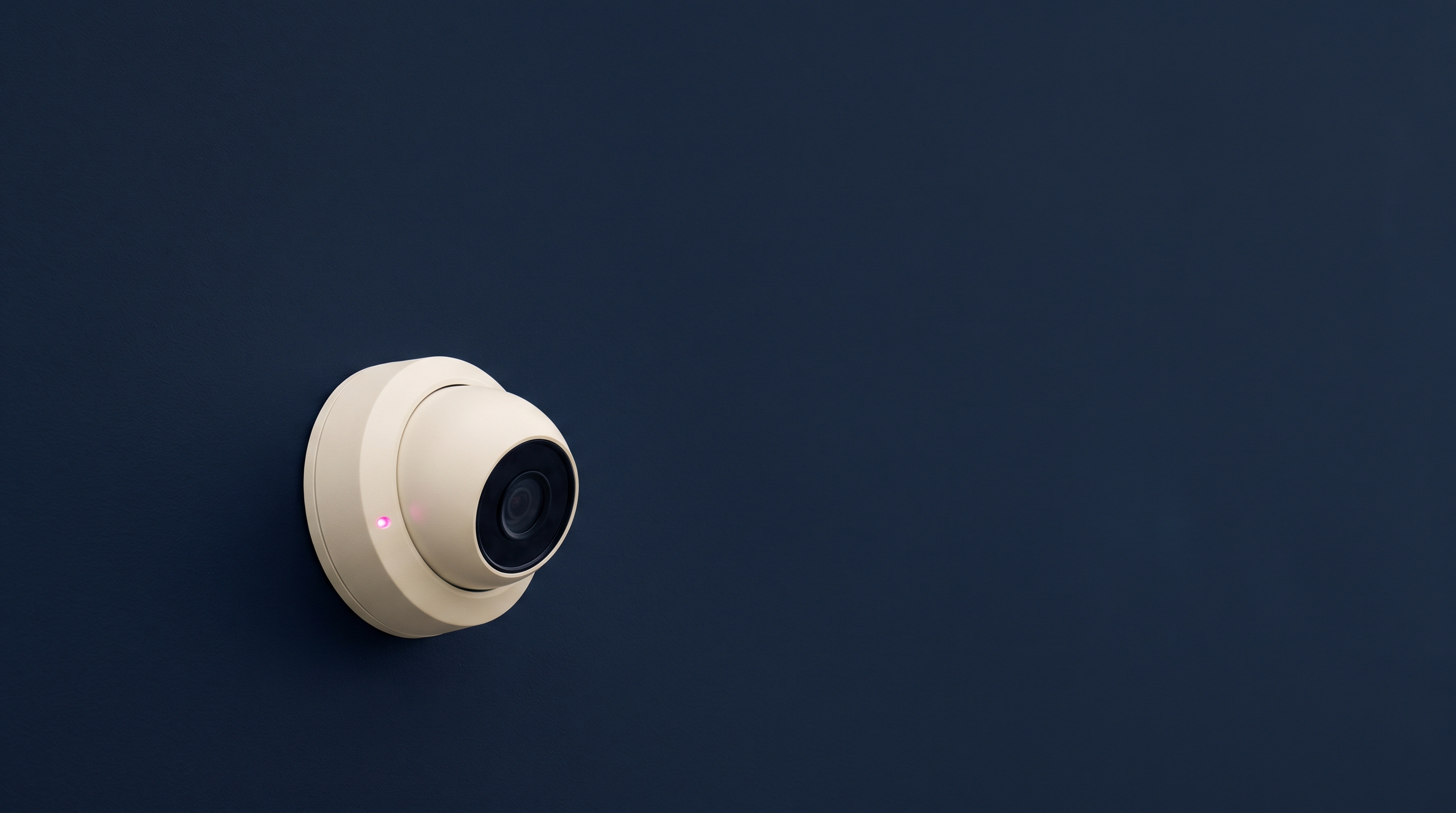 A security camera mounted high on a wall, angled away from the room and pointed instead at a blank wall, rendered in navy and cream tones with a single pink indicator light.