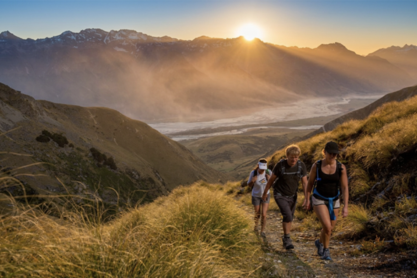 3 people walk up a mountain track with the sun behind them