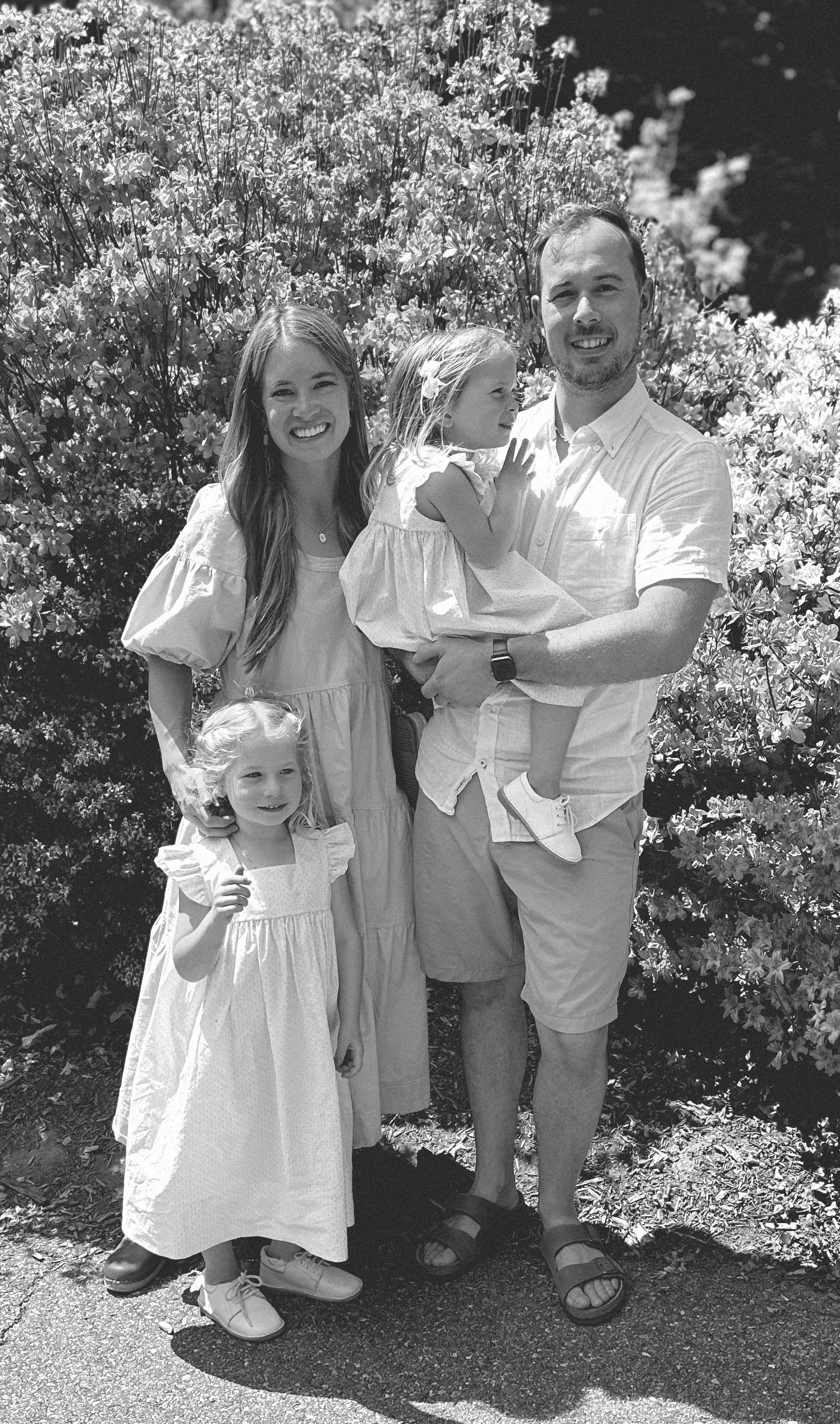 Smiling family of four outdoors with two young daughters in light dresses in front of a flowering bush.