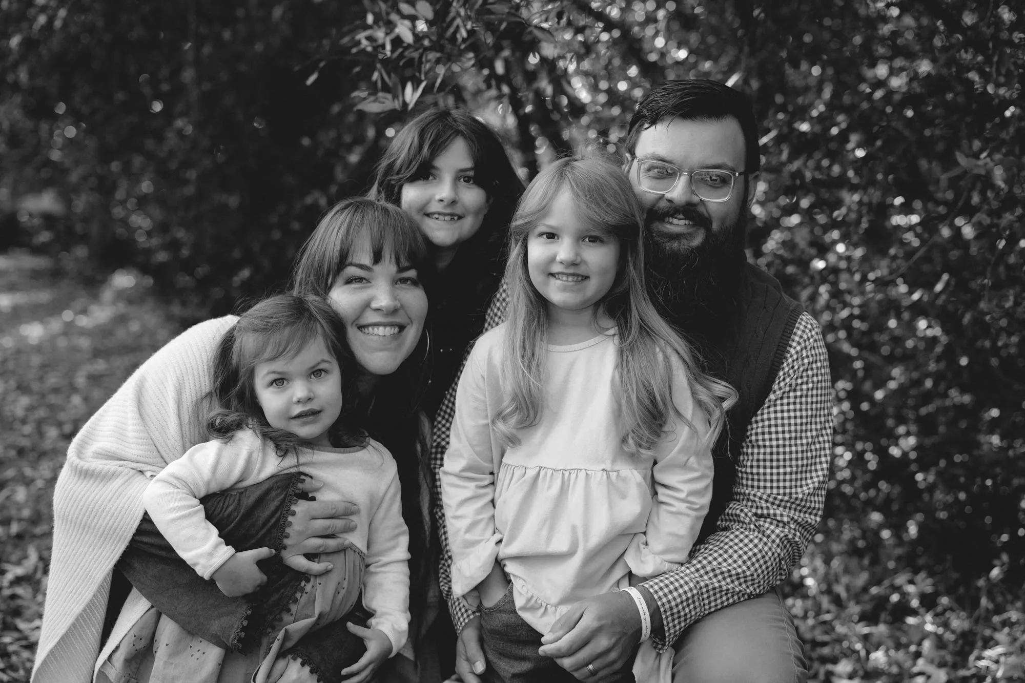 Black and white photo of a smiling family with a bearded man, woman, and three young girls outdoors.