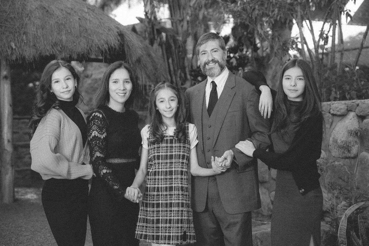Family of five posing outdoors with greenery and stone wall in the background, smiling at the camera.
