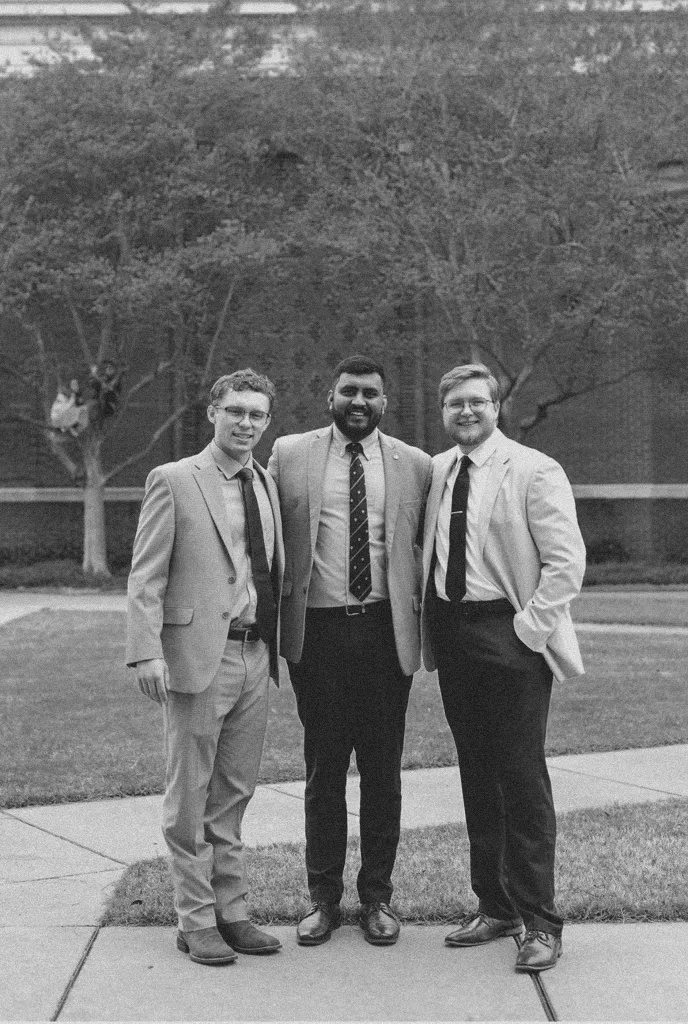 Three men in suits smiling and standing close together outside on a pathway near grass and trees.