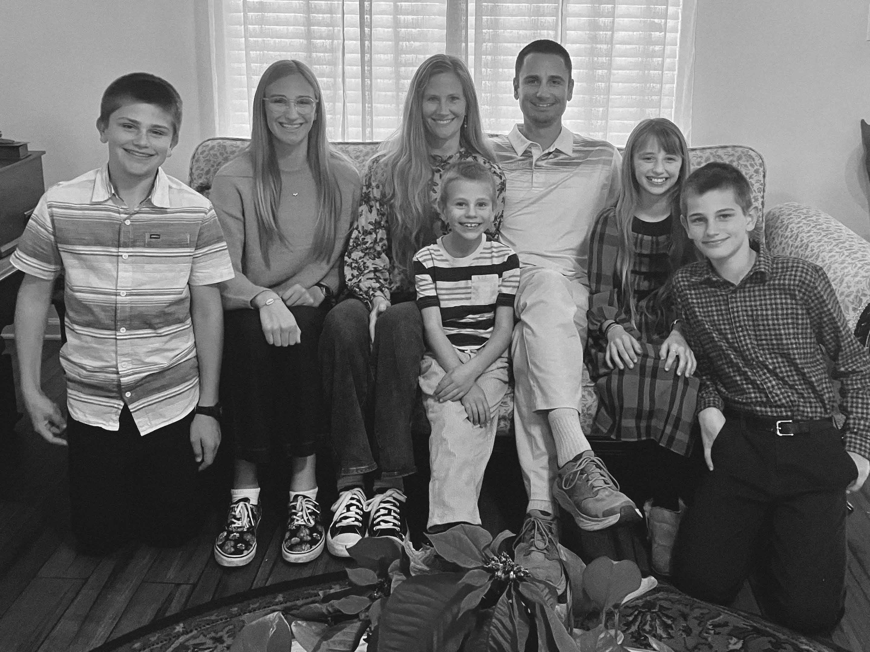 Seven smiling family members seated and kneeling in front of a window with blinds indoors.