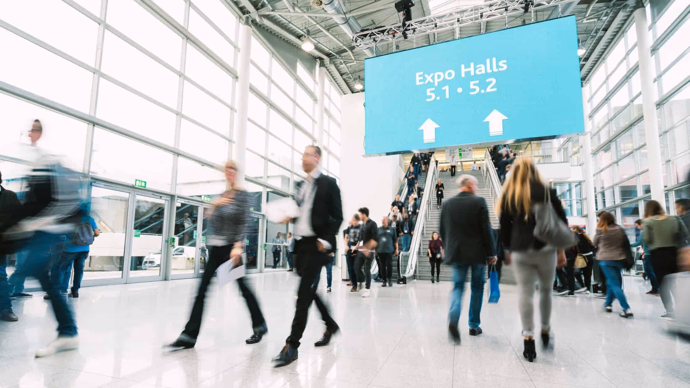 People walk through a busy convention center lobby with a large sign overhead directing attendees to Expo Halls 5.1 and 5.2.