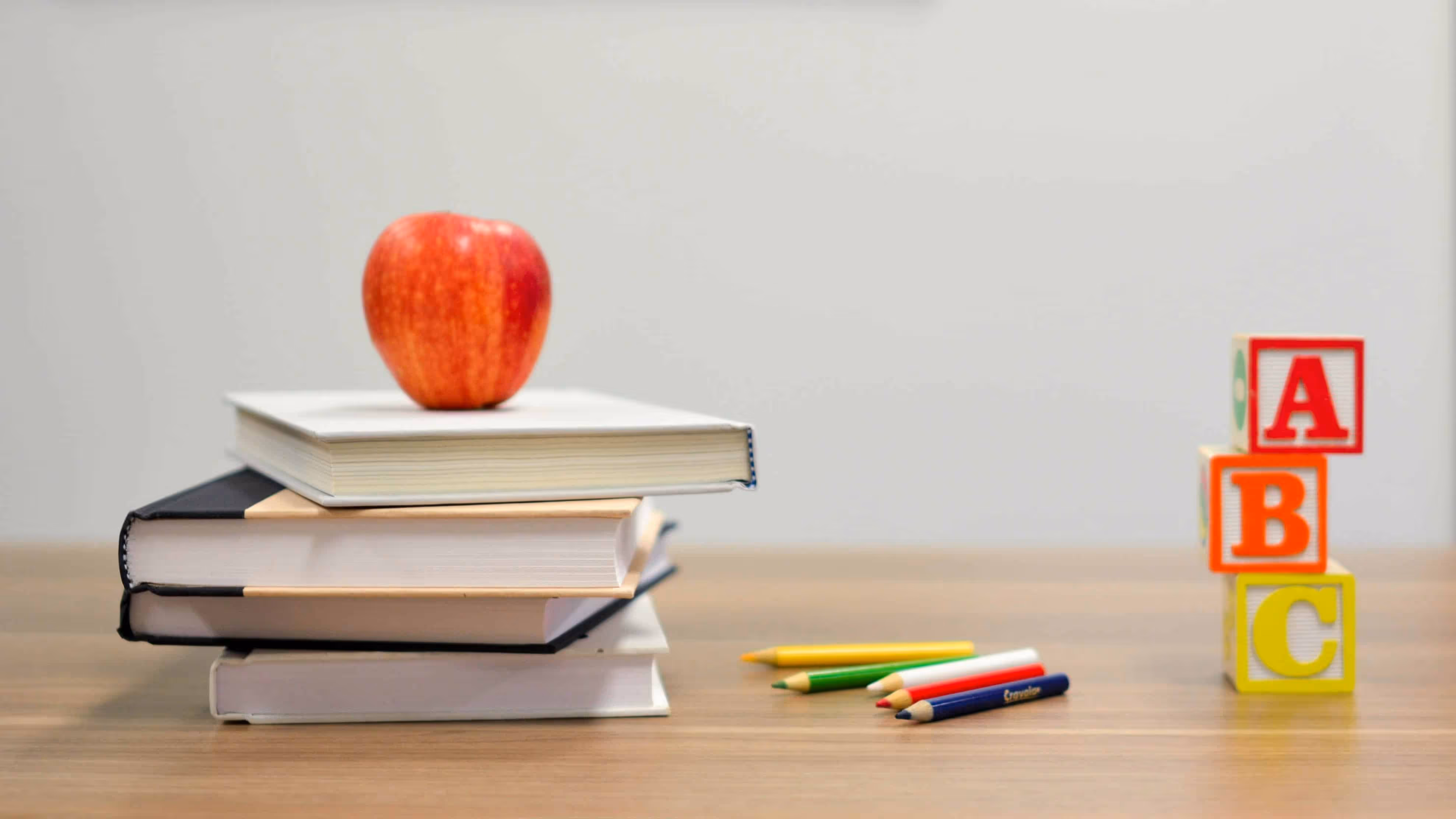 A red apple on top of a stack of books beside colored pencils and wooden alphabet blocks spelling ABC on a wooden desk.