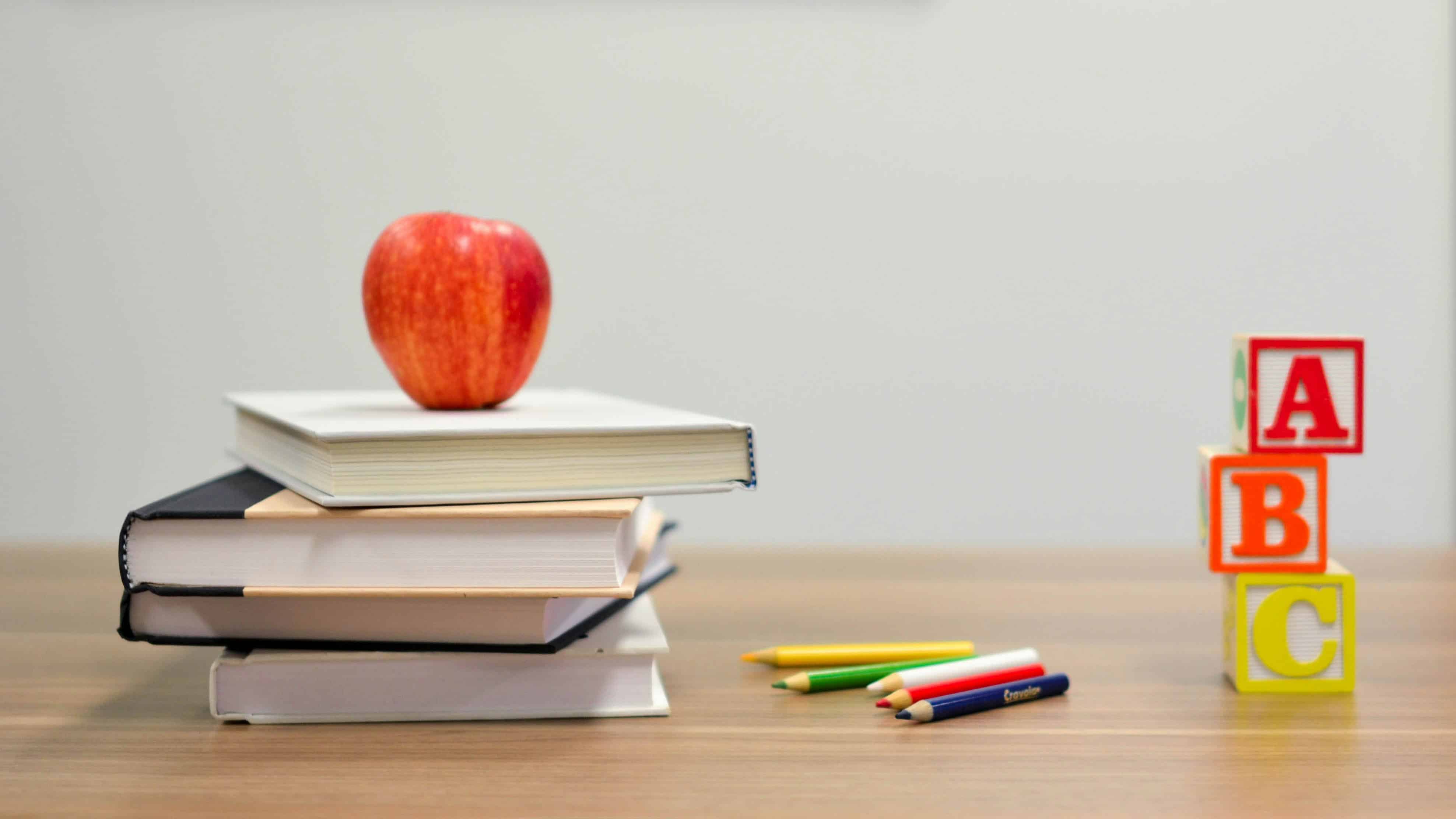 A red apple on top of a stack of books beside colored pencils and wooden alphabet blocks spelling ABC on a wooden desk.