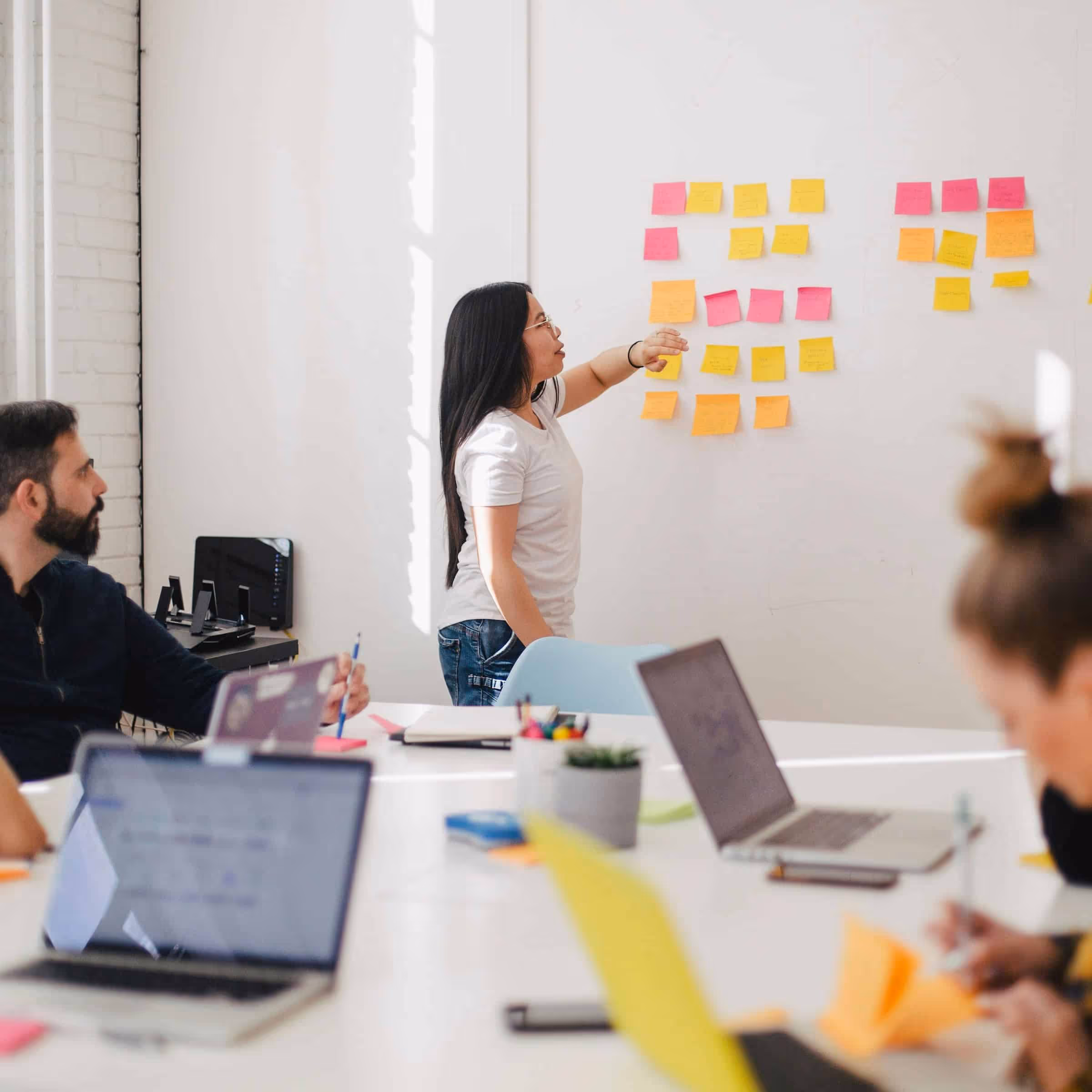 Team collaborating in a bright office, with a woman leading a brainstorming session by organizing sticky notes on a wall, while colleagues focus on laptops and discussions around a table.