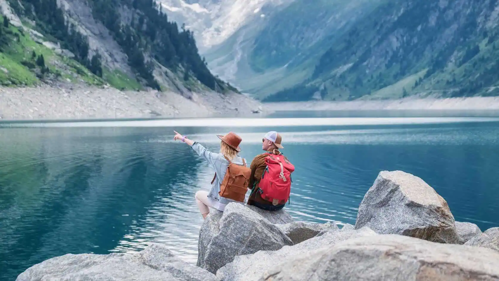 Two hikers with backpacks sitting on rocks by a mountain lake, one pointing toward the view