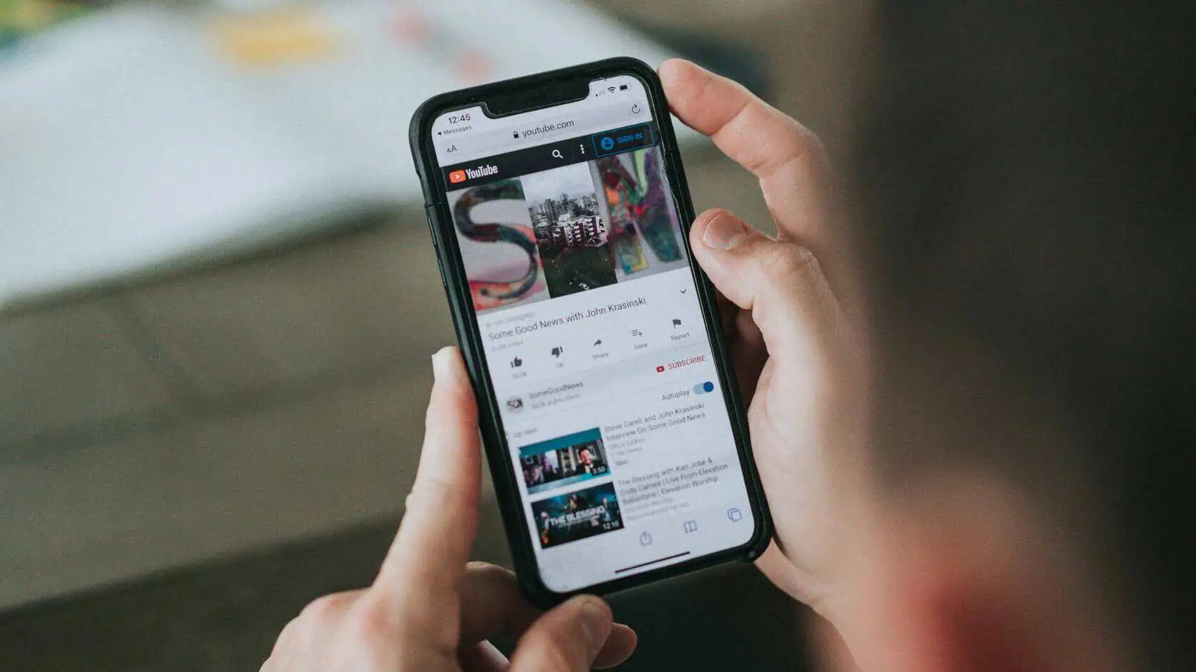 A person holds a smartphone displaying a YouTube video titled “Some Good News with John Krasinski.” The screen shows the colorful SGN thumbnail, video controls, and related videos below. The background includes a blurred tabletop with papers and a candle.