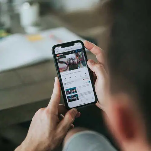 A person holds a smartphone displaying a YouTube video titled “Some Good News with John Krasinski.” The screen shows the colorful SGN thumbnail, video controls, and related videos below. The background includes a blurred tabletop with papers and a candle.