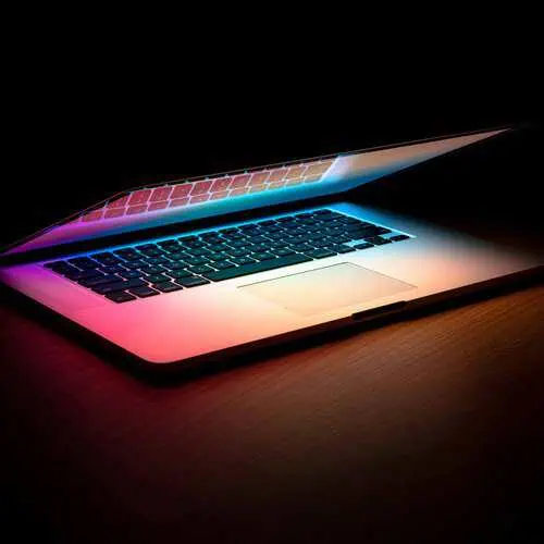 An open laptop on a dark wooden desk, lit with colorful pink and blue light against a black background.