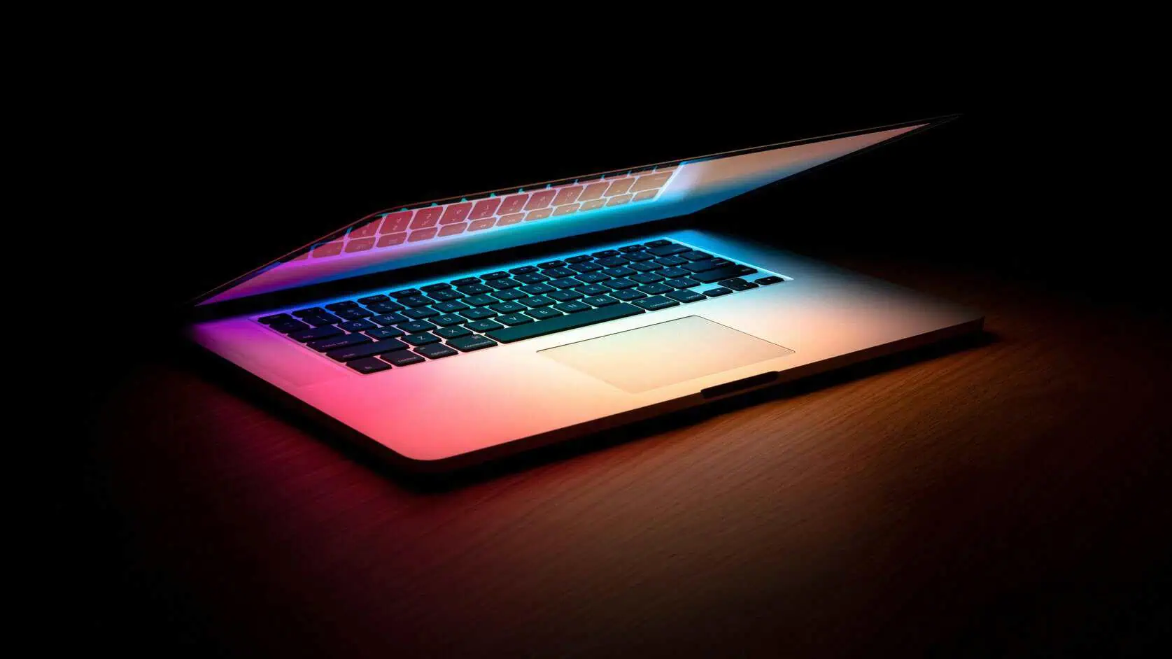 An open laptop on a dark wooden desk, lit with colorful pink and blue light against a black background.
