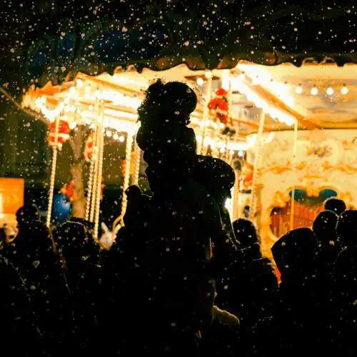 Silhouettes of a crowd, including an adult carrying a child on their shoulders, standing in falling snow in front of a brightly lit carousel at night.