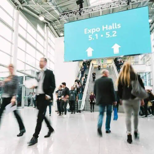 People walk through a busy convention center lobby with a large sign overhead directing attendees to Expo Halls 5.1 and 5.2.