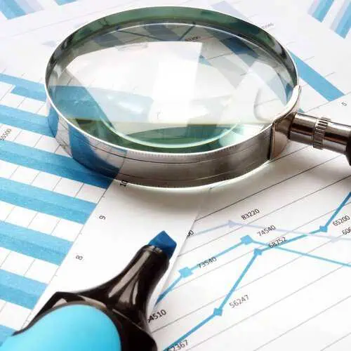 A silver magnifying glass is positioned over blue and white line graphs and bar charts displaying financial data. A blue marker rests near the charts, suggesting analysis.