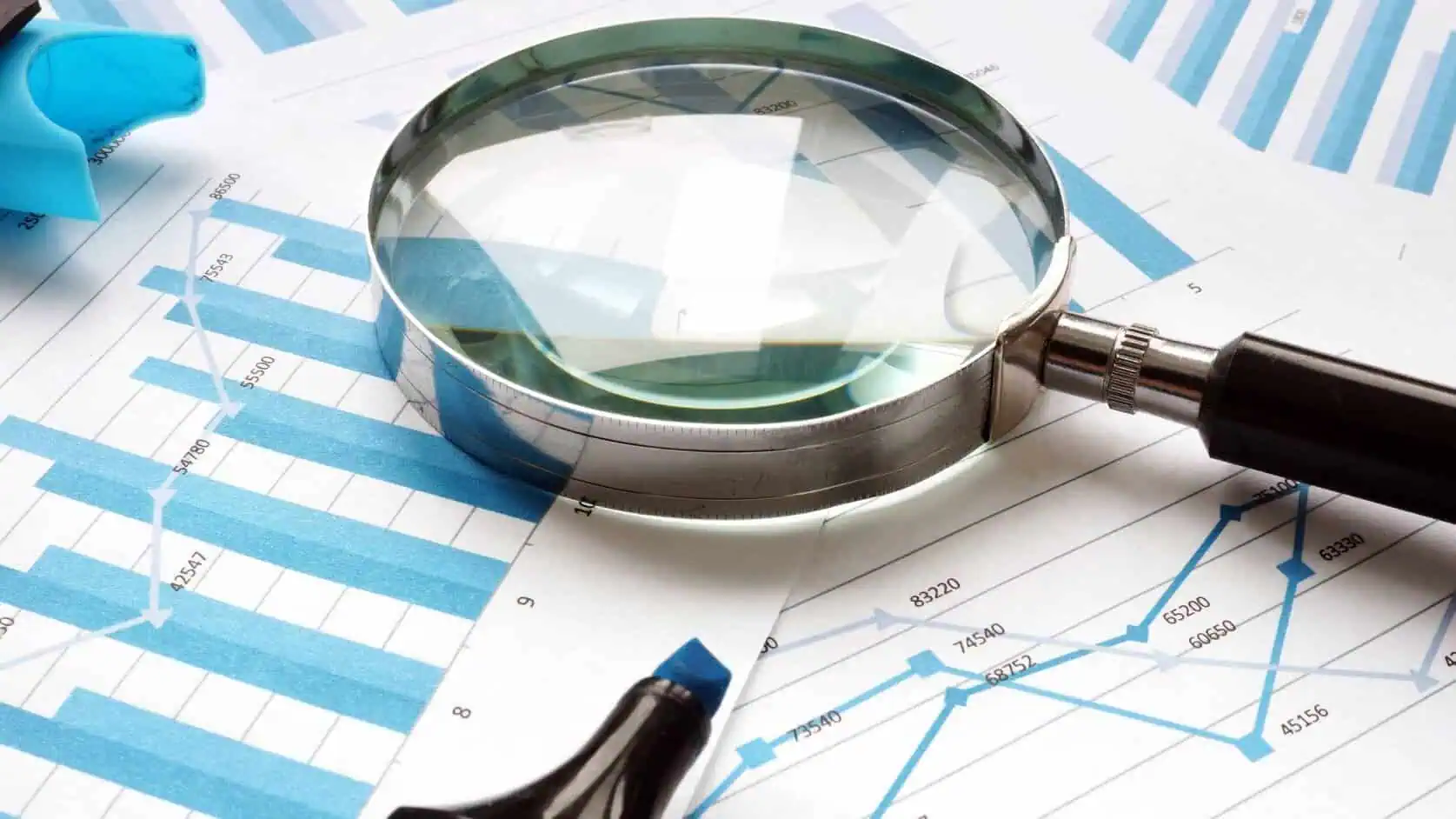 A silver magnifying glass is positioned over blue and white line graphs and bar charts displaying financial data. A blue marker rests near the charts, suggesting analysis.