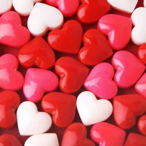 A close-up image of glossy, heart-shaped candies in red, pink, and white, arranged tightly together. The candies have a shiny, smooth surface, reflecting light.