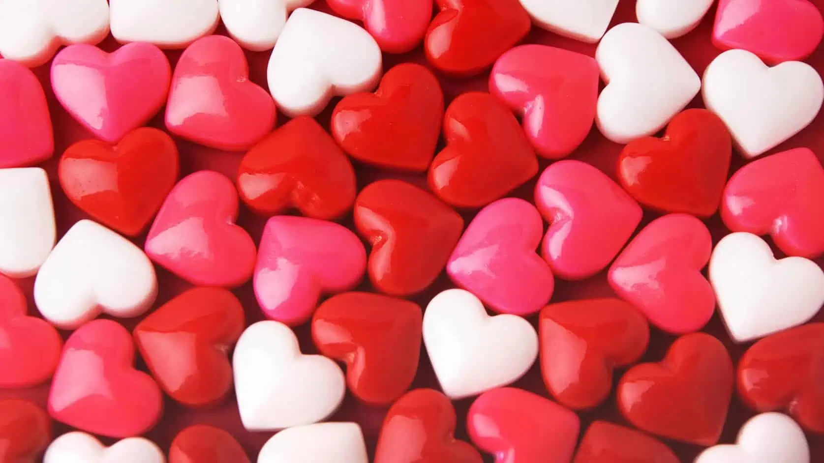 A close-up image of glossy, heart-shaped candies in red, pink, and white, arranged tightly together. The candies have a shiny, smooth surface, reflecting light.