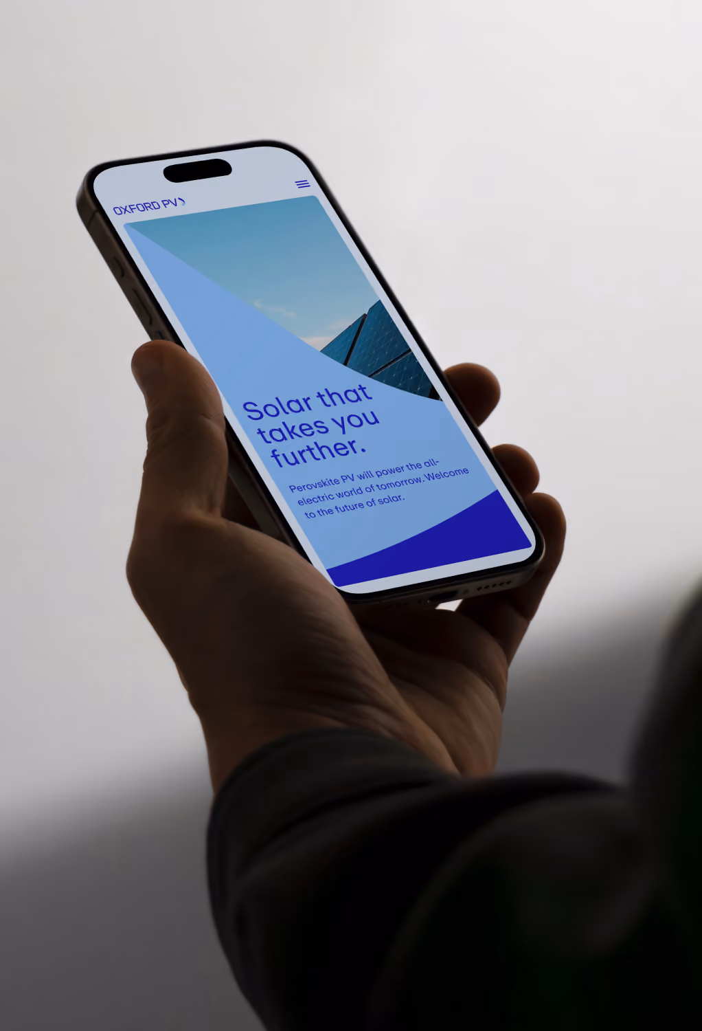 Hand holding phone showing Oxford PV website with solar energy message and solar panel image.