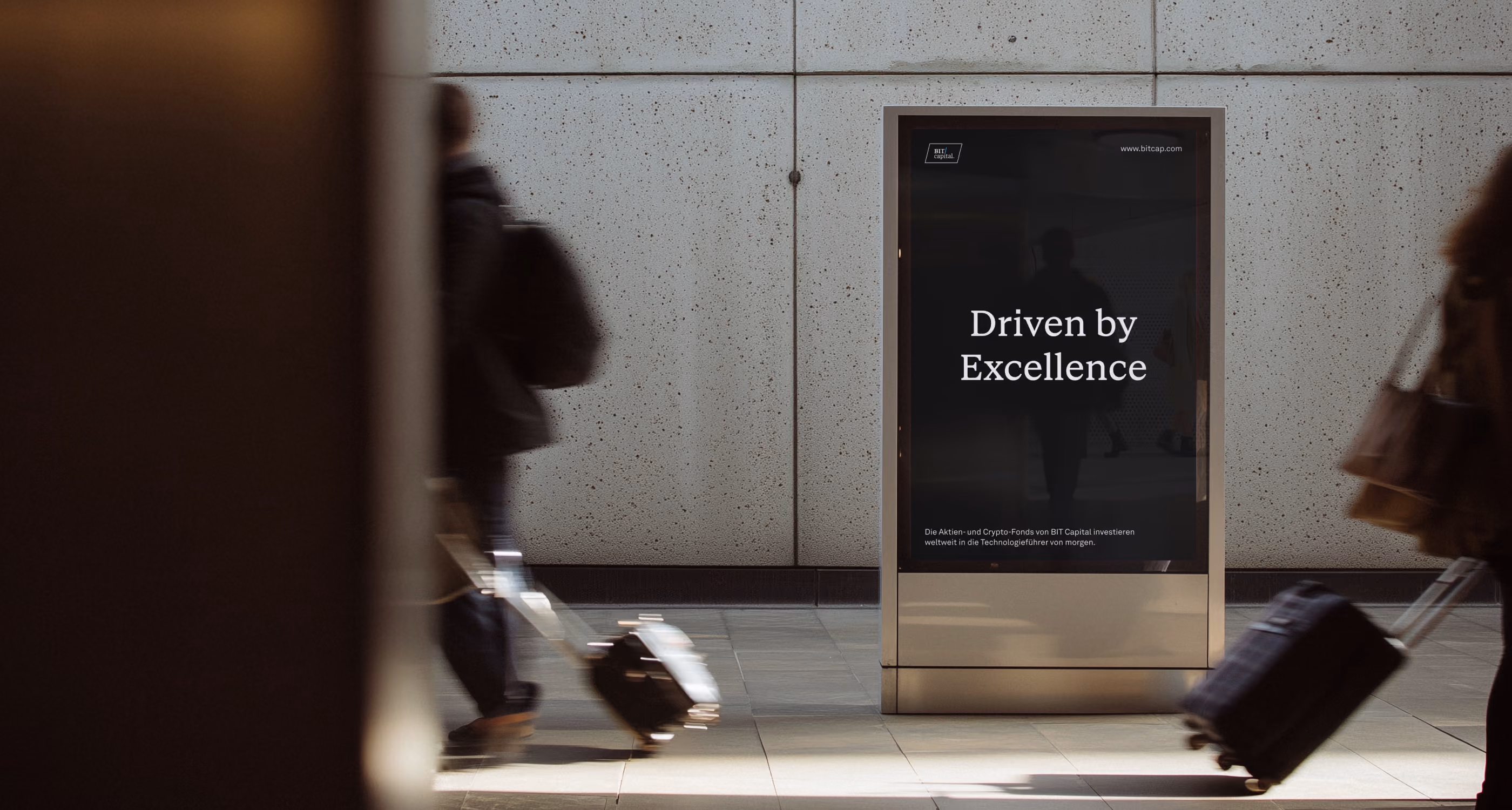Digital sign at station with text "Driven by Excellence" as blurred travelers pass by with suitcases.
