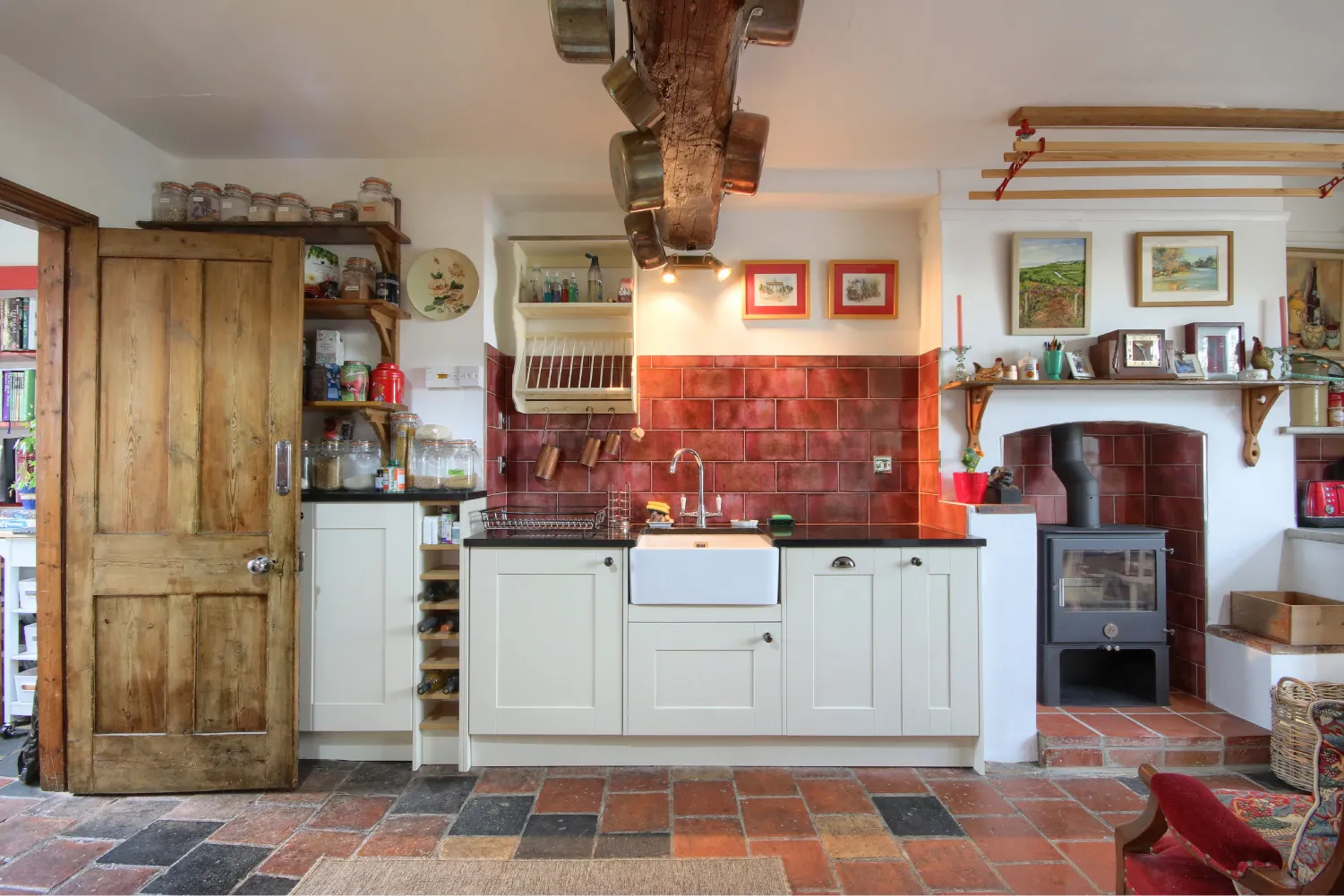 Cozy kitchen with white cabinets, farmhouse sink, red tiled backsplash, hanging copper pots from wooden ceiling beam, and a wood-burning stove set in a tiled alcove.