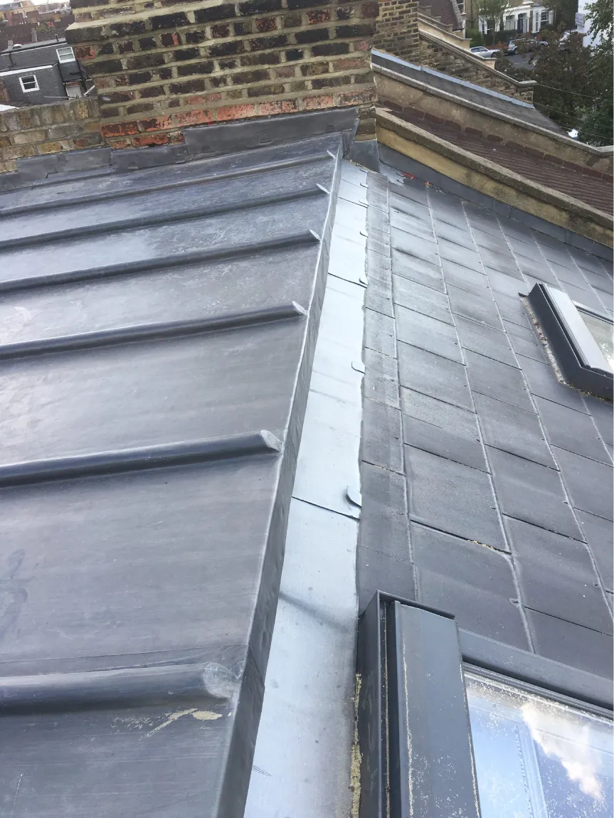 Close-up of a roof corner with metal flashing between a standing seam metal roof and dark slate tiles next to a brick chimney.