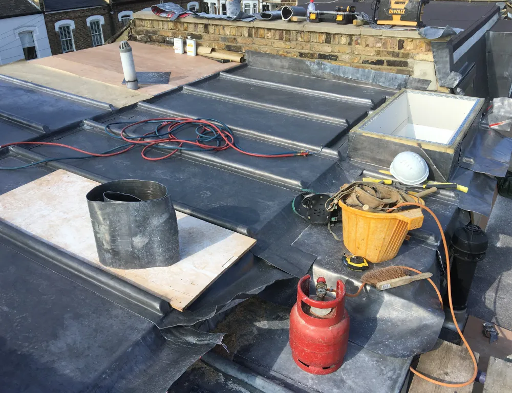 Roof under construction with black waterproof membrane, a red gas cylinder, coiled cables, a yellow bucket with tools, a white hard hat, and a brick chimney.
