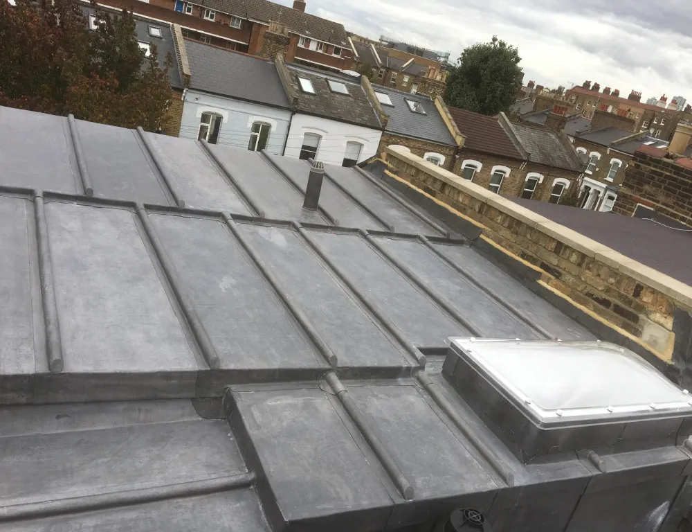 Flat grey metal roof with a small chimney pipe and a rectangular skylight, overlooking residential buildings with pitched roofs.
