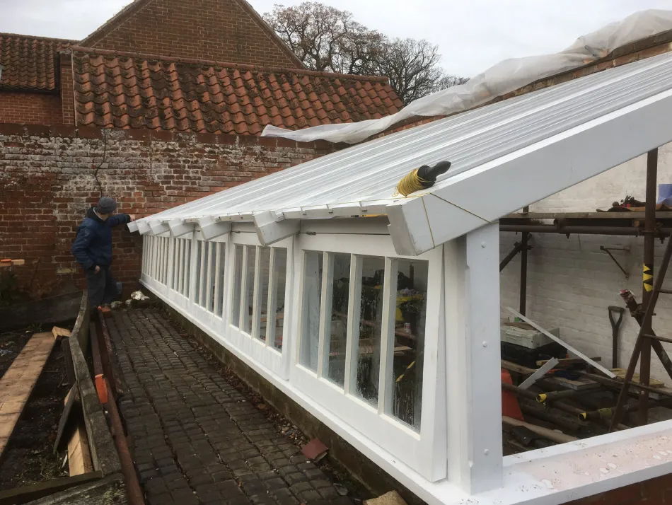 Person working on a newly constructed white greenhouse with slanted transparent roof panels beside a brick wall.