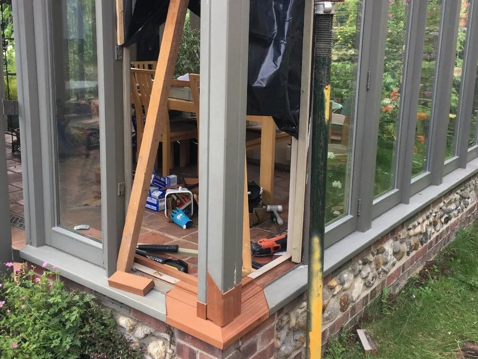 Close-up of corner of a glasshouse with gray window frames and stone base, showing wooden supports and tools inside during construction or repair.