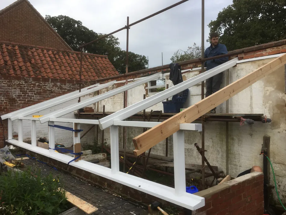 Construction site showing wooden and white painted beams forming a framework attached to a brick wall with scaffolding and two men working.