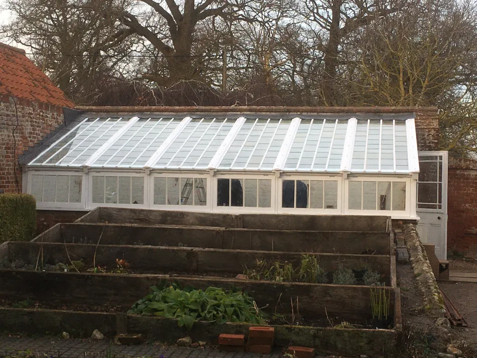 A white-framed glasshouse with a pitched roof next to a brick wall, with several tiered raised garden beds in the foreground.
