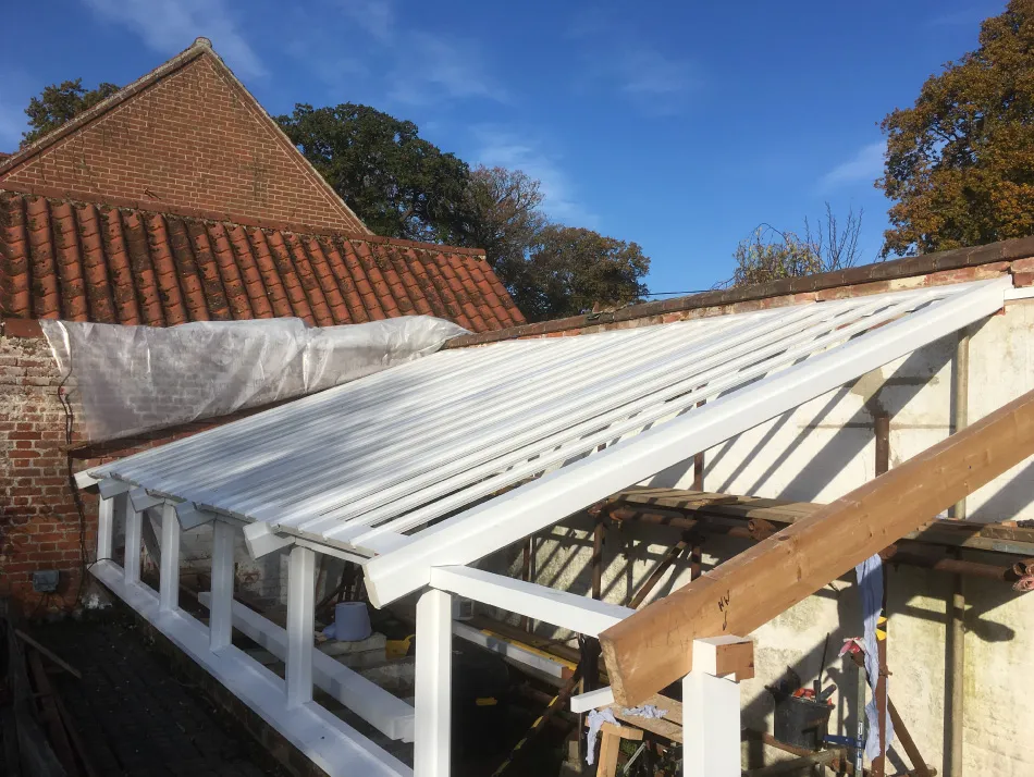 Partially constructed white wooden greenhouse frame with roof slats, set against a brick wall and under a blue sky.