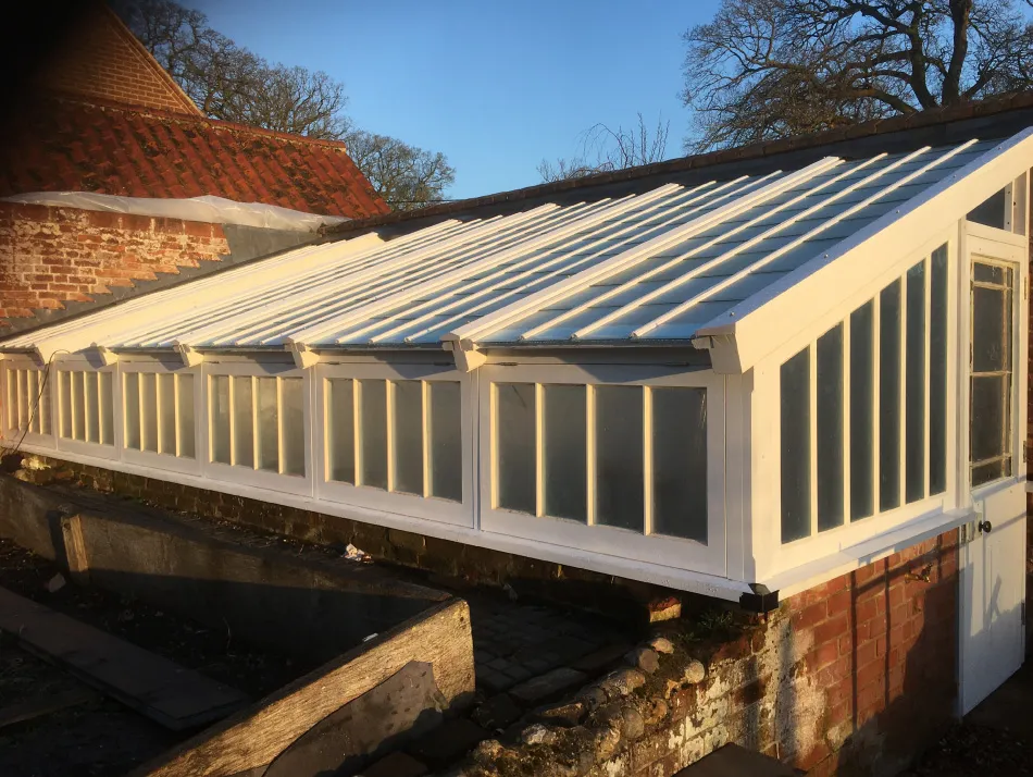 White-framed greenhouse with slanted roof panels attached to a brick wall, under a clear blue sky.